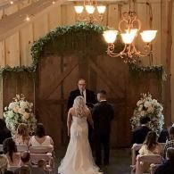 Bride and groom stand before an officiant in a church aisle under floral arch décor