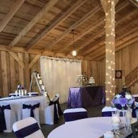Rustic barn venue set for an event, with round tables, purple accents, and a dessert display.