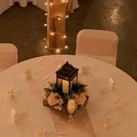 Round table with white cloth and floral lantern centerpiece at a warmly lit event.