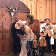 Cowboy in tan hat embraces his new bride in a wooden hall, with people standing nearby.