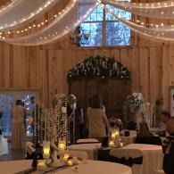 Rustic barn reception with draped string lights, round tables, and floral centerpieces under a high window