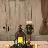 Rustic table centerpiece with lantern, candles, flowers, and white wooden cabinet backdrop