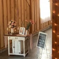 Rustic entryway with a white table, flowers, framed photo, and chalkboard sign beside string lights