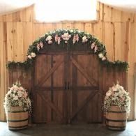 Rustic barn doors decorated with pink and white floral arrangements and greenery, flanked by flower-filled barrels.