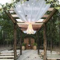 Wooden archway draped with white fabric and greenery, leading to an outdoor wedding aisle in the woods