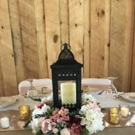 Decorated table centerpiece with a black lantern, pink and white flowers, and gold candle holders against a wood wall