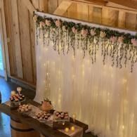 Rustic wood backdrop with hanging flowers, warm fairy lights, and a decorated dessert table