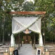 Outdoor wedding arch with white drapes and greenery under trees, lined walkway leading to a chandelier.