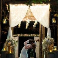 Bride and groom kissing under a glowing floral arch at an outdoor wedding ceremony