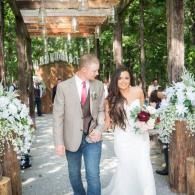 Bride and groom walking down a flower-lined outdoor aisle, smiling and holding hands.