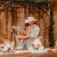 Couple feeding each other at a rustic wedding cake table with white floral decor