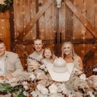 Wedding party posed in front of rustic barn doors, holding a white hat amid floral arrangements.