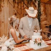 Couple in western attire cutting a white wedding cake at a rustic barn reception