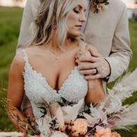 Bride in white lace dress holding bouquet, with groom’s hand on her shoulder outdoors at sunset