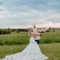 Bride in a white lace gown holds a bouquet in a grassy field under a cloudy sky.