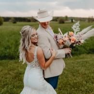 Bride hugging partner in a white hat while holding a bouquet in a green field at sunset