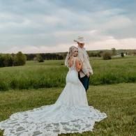 Bride and groom standing in a grassy field at sunset, with the bride’s long lace train spread behind her