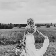 Bride in a white dress walking through a field, holding a bouquet and lifting her train