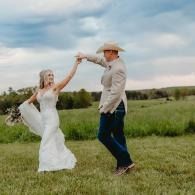 Couple dancing in a grassy field under a cloudy sky, the woman in a white dress and the man in a cowboy hat