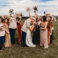 Bride and friends posing in a grassy field, laughing and holding bouquets under a cloudy sky