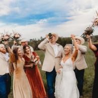 Wedding party cheering outdoors, with bride in white and bridesmaids in rust dresses raising drinks under a blue sky