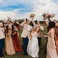 Wedding party celebrating outdoors, raising hats and bouquets in a grassy field under a cloudy sky
