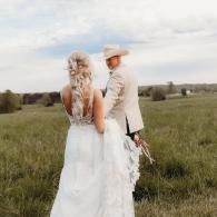 Bride and groom walking through a grassy field, holding hands under a cloudy sky.