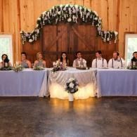 Wedding party seated at a long head table under a floral arch in a rustic wood-paneled hall