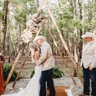 Bride and groom kissing under a rustic wedding arch in a wooded outdoor ceremony