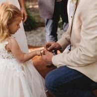 A child in a white dress is getting a ring placed on her finger by an adult during an outdoor ceremony.