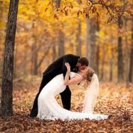 Couple in wedding attire kissing in an autumn forest with golden leaves.