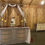 Rustic event table with draped white fabric, candles, and tiered cake in a wooden barn setting