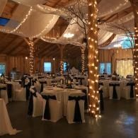 Elegant banquet hall with round tables, black-and-white chairs, and string lights draped from wooden beams