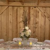 Rustic wedding table centerpiece with white flowers, candles, and branches against a wooden barn wall