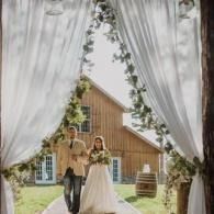 Couple walking through white drapes toward a rustic barn wedding venue, framed by greenery.