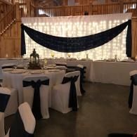 Decorated banquet hall with round tables, white chair covers, and dark blue drapery against a lit backdrop