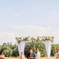 Outdoor wedding ceremony under a white floral arch in a field, with guests and bridal party nearby