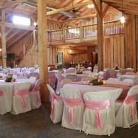Rustic barn-style banquet hall with round tables and white chairs tied with pink sashes