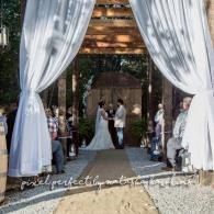 Bride and groom stand under a floral arch at an outdoor wedding aisle with white drapes.