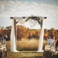 Decorated white wedding arch with draped fabric and greenery in an outdoor field at sunset
