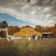 Rustic yellow barn at an outdoor gathering with people, tents, and autumn trees under a cloudy sky
