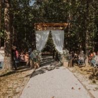 Outdoor wedding aisle with white drapery, greenery arch, and guests seated on both sides
