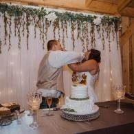Couple feeding cake at a rustic wedding dessert table with greenery backdrop