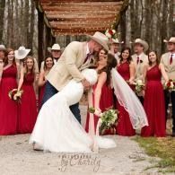 Bride and groom kiss during outdoor wedding ceremony, with bridesmaids in red dresses behind them.