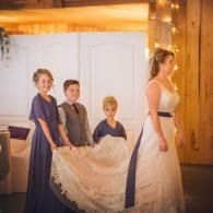 Three children carry a smiling woman’s dress train in a warmly lit wooden interior.