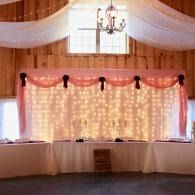 Decorated banquet hall with white drapes, pink lights, and long covered tables under a chandelier