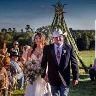 Bride and groom walk down an outdoor aisle at a rustic wedding with guests seated nearby.