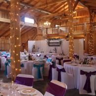Rustic barn venue with white tables, purple chair sashes, and string lights.