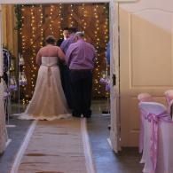 Couple standing at an indoor wedding arch with string lights and pink chair sashes