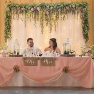 Wedding couple seated at a decorated head table with pink draping and hanging greenery backdrop